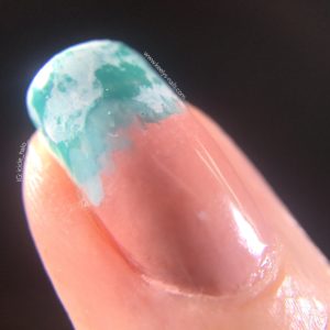 A macro photo of right ring finger, with a black background. The tip of the nail has a sea design using turquoise and white nail polish to imitate waves on a beach.